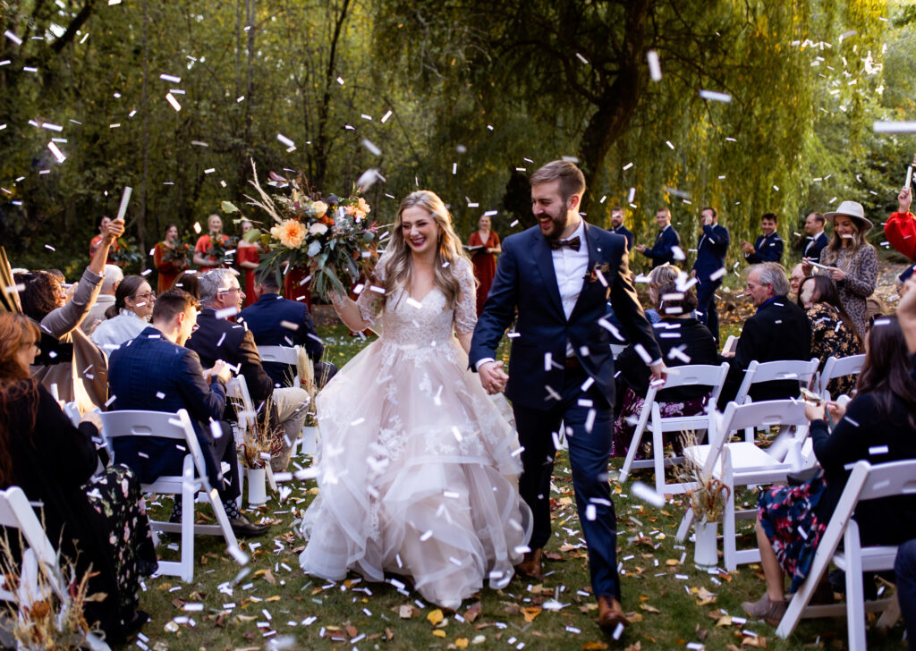 A newlywed couple walks hand in hand through a joyful confetti exit during their Portland wedding, captured by Wild Elopements Photography. The bride, in a romantic lace gown, holds a lush bouquet of fall florals, while the groom beams beside her in a navy suit. Guests cheer under the golden light of an outdoor woodland setting, creating a vibrant and celebratory moment.